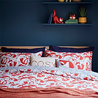 A double bed with a fox print duvet set and matching accent cushions and throws, with a wall shelf above the headboard with fox ornaments and books stacked on top