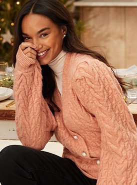 A smiling woman wearing a cream turtleneck, pink knitted cardigan and black trousers. 