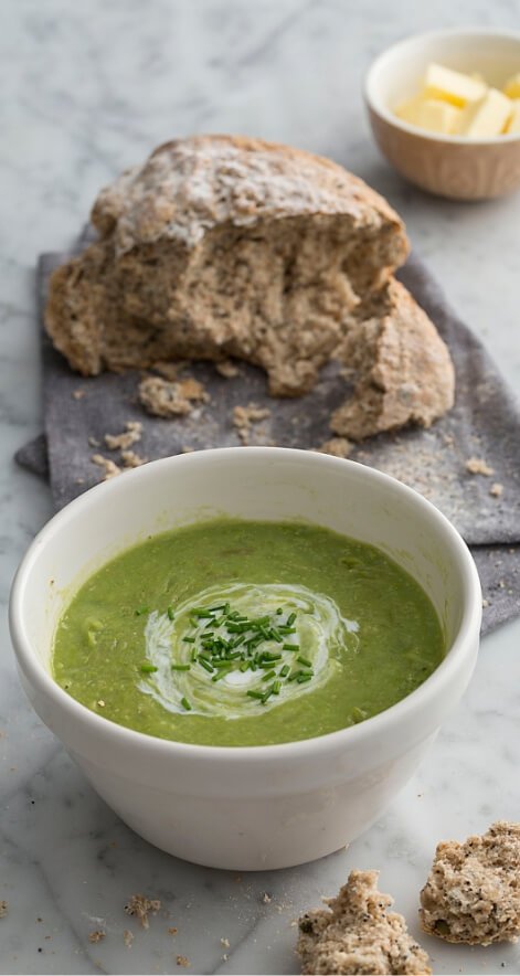 A bowl of green soup, a loaf of bread and a small bowl of butter cubes.