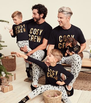 Two men and two children and dog sit together smiling wearing matching black cosy crew family Christmas pyjama set. 