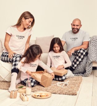 Woman and man sit on sofa smiling at two young girls sitting on the floor opening gifts, wearing cream and black Christmas family matching lounging pyjamas set.