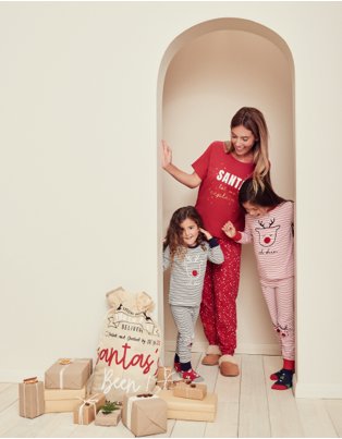 Woman and two young girls stand smiling in doorway wearing Christmas pyjamas looking at Santa sack and pile of wrapped presents.