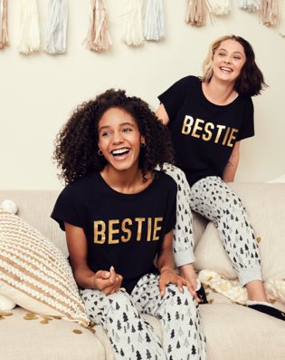 Two women sit smiling on cream sofa wearing black, white and gold bestie slogan family Christmas pyjamas.