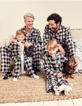 Two men, two young boys and dog sit smiling wearing black and white check print family Christmas pyjamas.