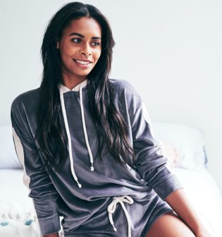 Woman poses on bed wearing grey velour hoodie and shorts.