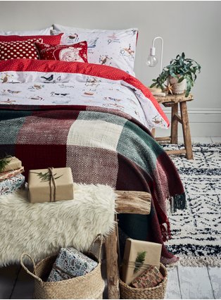 Double bed with wooden table, white lamp and plant to right hand site with multi 12 days of Christmas reversible duvet set, check print throw and wrapped presents at the bottom of the bed. 