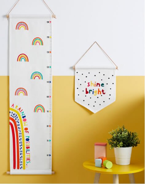 A yellow side table with a potted plant and toys in front of a yellow and white wall with a rainbow height chart and a 'Shine Bright' wall hanging.