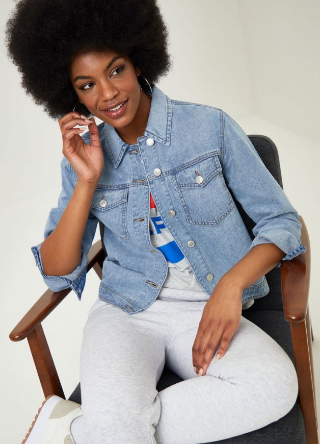 A woman sitting in a chair wearing white trousers and a light wash denim jacket.