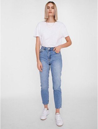 Woman poses with hand in pocket wearing white t-shirt tucked into NOISY MAY blue mid wash high waist mom jeans and white trainers.