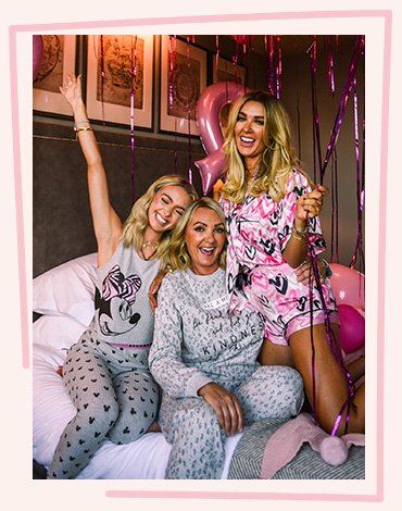 Three smiling women huddle together on a large white bed wearing pink and grey pyjamas from the Tickled Pink collection with pink balloons and streamers in the background.