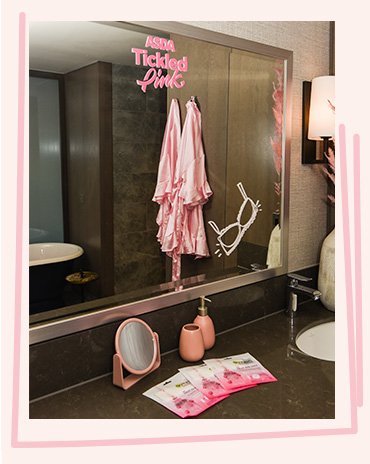 Grey hotel bathroom features large mirror, pink silky robe, ASDA Tickled Pink wall stickers, pink soap dispenser, tumbler, small mirror and three pink face masks.
