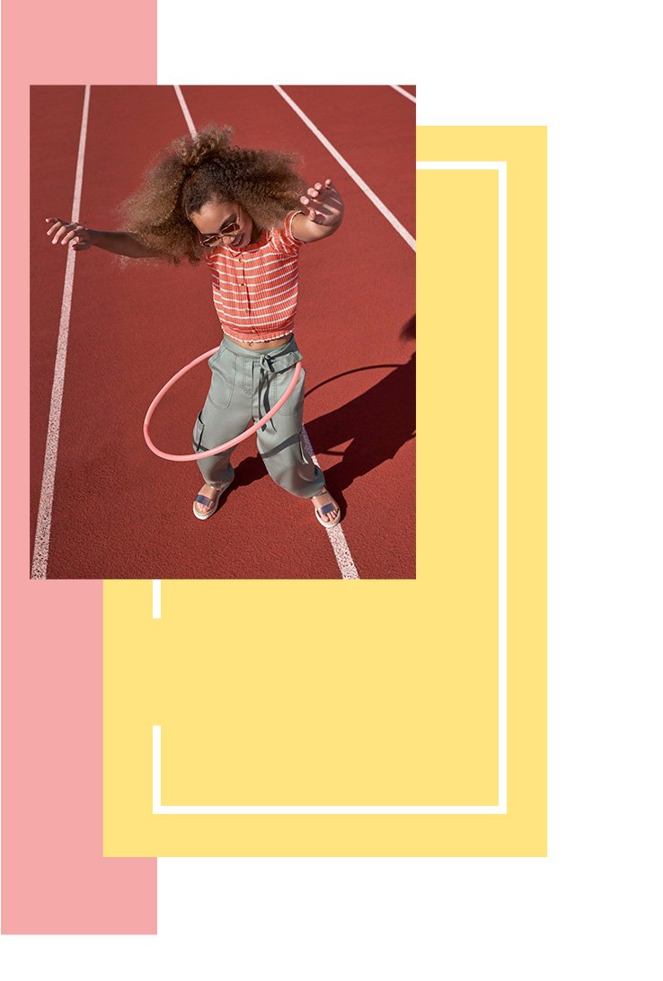 Girl hula-hooping on a sports court wearing a red striped button up top, grey cargo trousers and metallic sandals