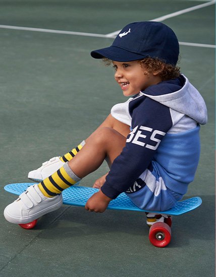 Boy sitting on a skateboard on a sports court in a navy slogan panel hoodie, matching shorts, a navy cap and white trainers