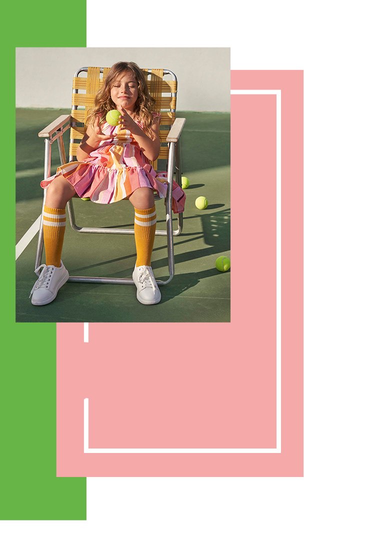 Girl sitting on a picnic chair on a sports court holding a tennis ball wearing a pink striped sleeveless dress, high orange socks and white trainers
