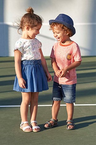 A girl and boy on a sports court, the girl wearing a white frill sleeve top, denim skirt and white sandals, the boy wearing a red lion logo T-shirt, denim shorts, a denim hat and sandals