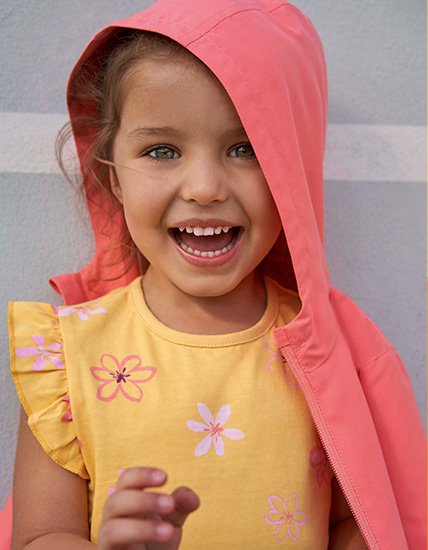 Girl smiling outside wearing an orange floral top and a pink jacket with the hood up