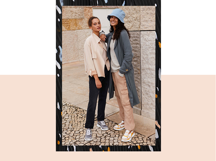 Two women outdoors, one wearing a nude jacket, black trousers and trainers, the other wearing a blue hat and grey longline coat over a jumper and pink trousers