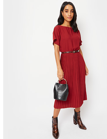 Woman wearing a dark red pleated midi dress with black shell buckle belt, black handbag and boots