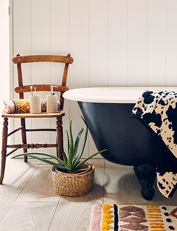 Bath with cow print towel, plant and wooden chair with bathroom accessories, and a bed with patterned cushions, plants and a cow print rug 