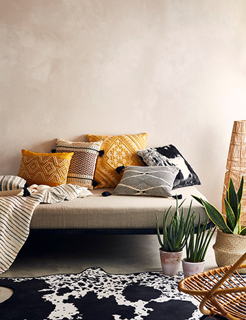 Bath with cow print towel, plant and wooden chair with bathroom accessories, and a bed with patterned cushions, plants and a cow print rug 