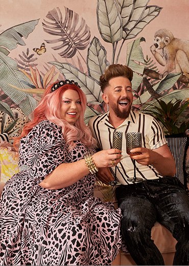 Woman smiles wearing animal print dress toasting glasses with man wearing cream and black striped short-sleeved shirt and jeans, gold-tone drinks trolly features assorted wine glasses, champagne flutes and tumblers.