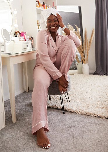 A woman sits on stool in dressing room wearing pink pyjamas with white towel on her head, cream room features sofa, multicoloured cushions with leaf and slogan wall art in the background.