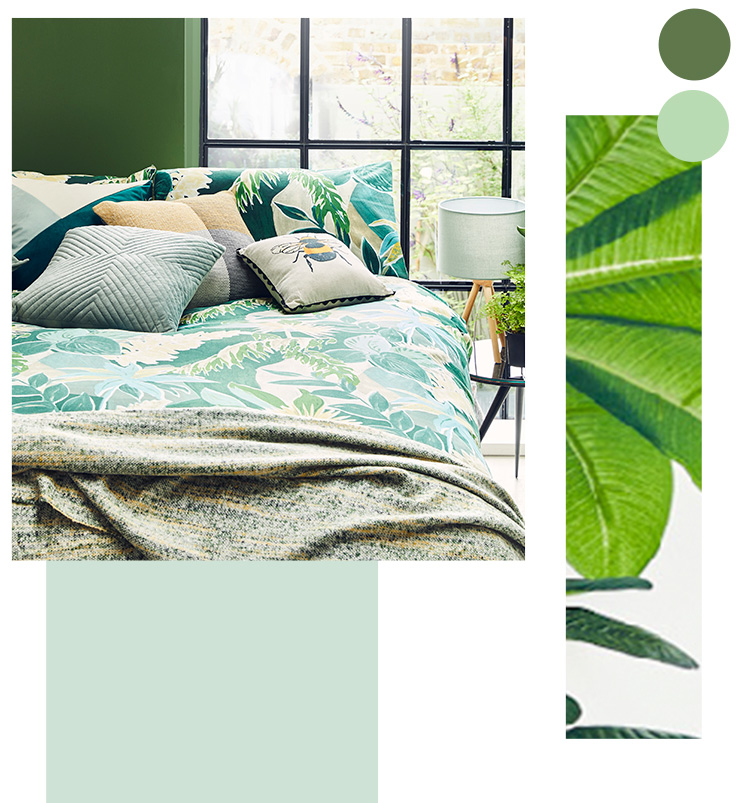 Double bed with green leaf print bedding, an assortment of cushions, a throw and a bedside table with an artificial plant