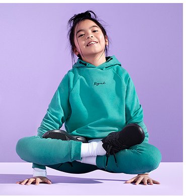 A girl doing a yoga pose wearing a teal hoodie and joggers outfit with white socks and black trainers.