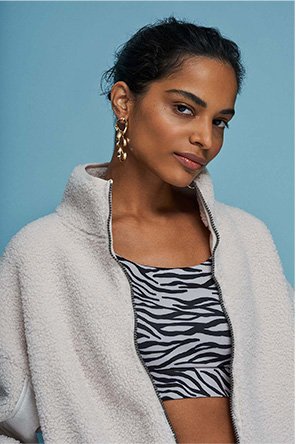 Woman poses in front of blue background wearing black and white zebra print crop top and cream high neck zip up jumper.