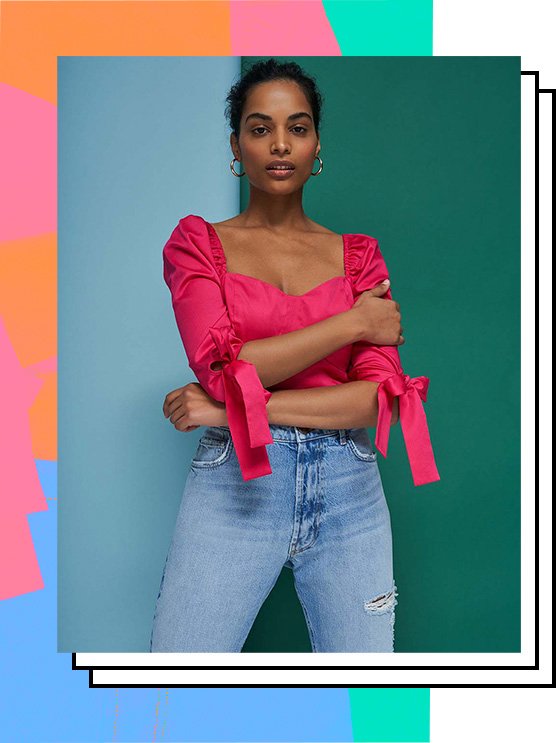 Woman poses in front of blue and green background with hands across torso wearing shocking pink ruffle tie sleeve top and distressed blue jeans.