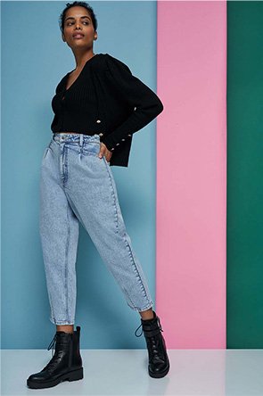 Woman poses with one foot forward in front of blue, pink and green background wearing black v-neck cami, black cardigan, acid wash mom jeans and black biker boots.