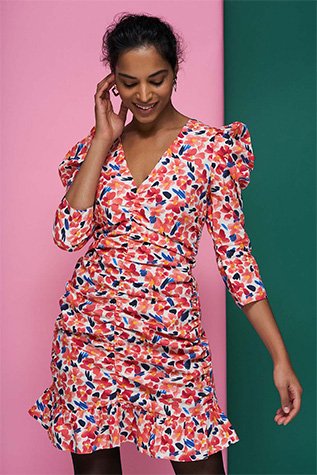 Woman smiles raising one hand to face in front of green and pink background wearing bright floral dress.