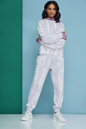 Woman poses with arms folded in front of green and blue background wearing purple hoodie, white coordinating loungewear top and trousers and white trainers.