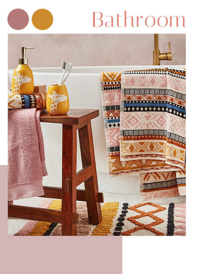 A bathroom with tribal patterned towels over a bath, a matching rug and a table with giraffe tumbler and dispenser