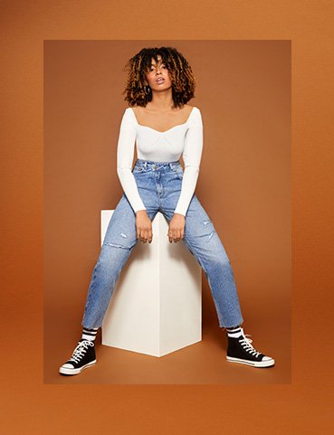 Phoenix Brown sits on white box wearing cream rib knit long sleeved bodysuit, blue light wash jeans and black canvas high top trainers.