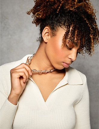 Close up of Phoenix Brown looking down wearing cream collared rib knit dress holding gold-effect necklace.
