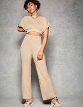 Phoenix Brown poses with hand on hip wearing beige co-ord knit crop top and wide leg trousers and brown trainers.