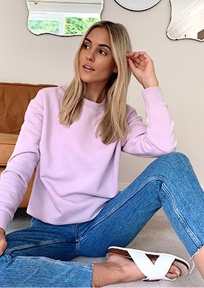 Woman sitting on the floor wearing a lilac crew jumper, jeans and white sandals