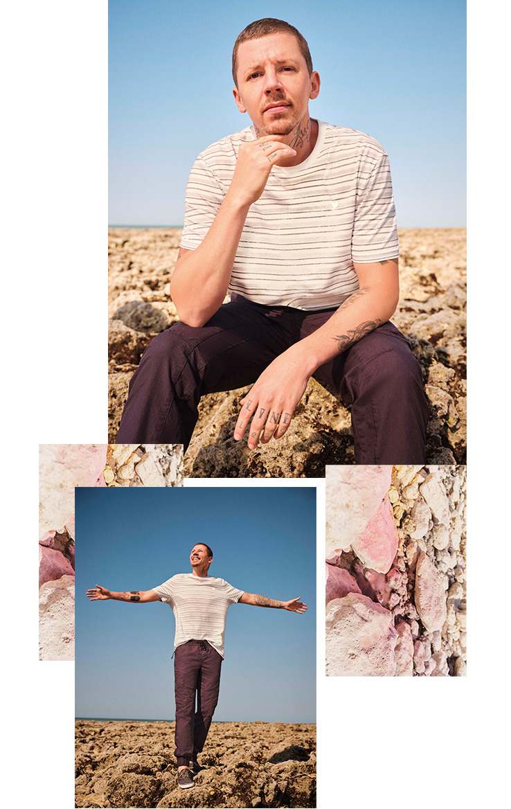 Professor Green wearing a white striped t-shirt and dark trousers on a rocky beach