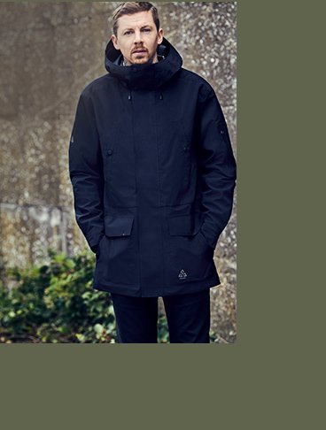 Professor Green stands with hands in pockets wearing navy coat and blue trousers with wall and greenery in the background.