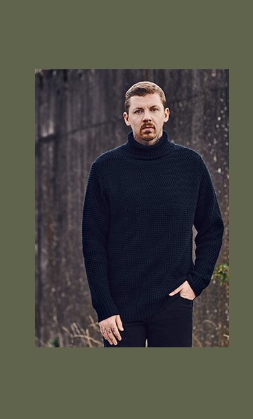 Professor Green stands with one hand in pocket wearing navy ribbed turtleneck and black trousers.