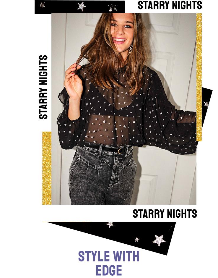 Brunette woman smiles and twirles hair wearing grey jeans with black belt and a black bralet with a sheer black and white star printed shirt.