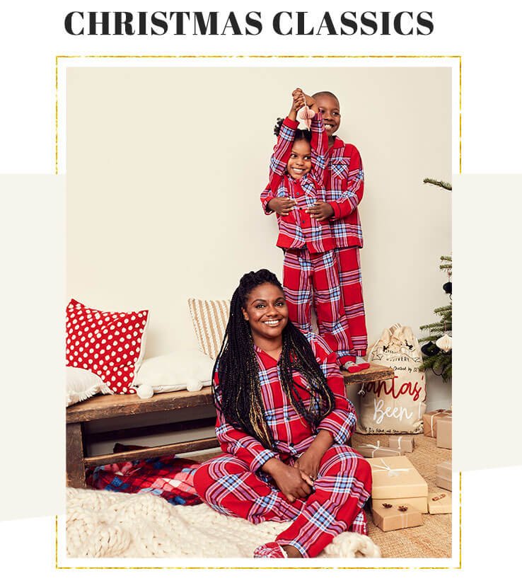 Woman sits crossed legged on the floor with a boy and a girl standing behind on a wooden bench all wearing red check print family matching Christmas pyjamas surrounded by wrapped gifts and a santa sack.