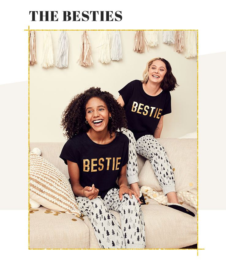 Two smiling women sit on a cream sofa surrounded by natural coloured scatter cushions both wearing black and gold bestie slogan family matching Christmas pyjamas.