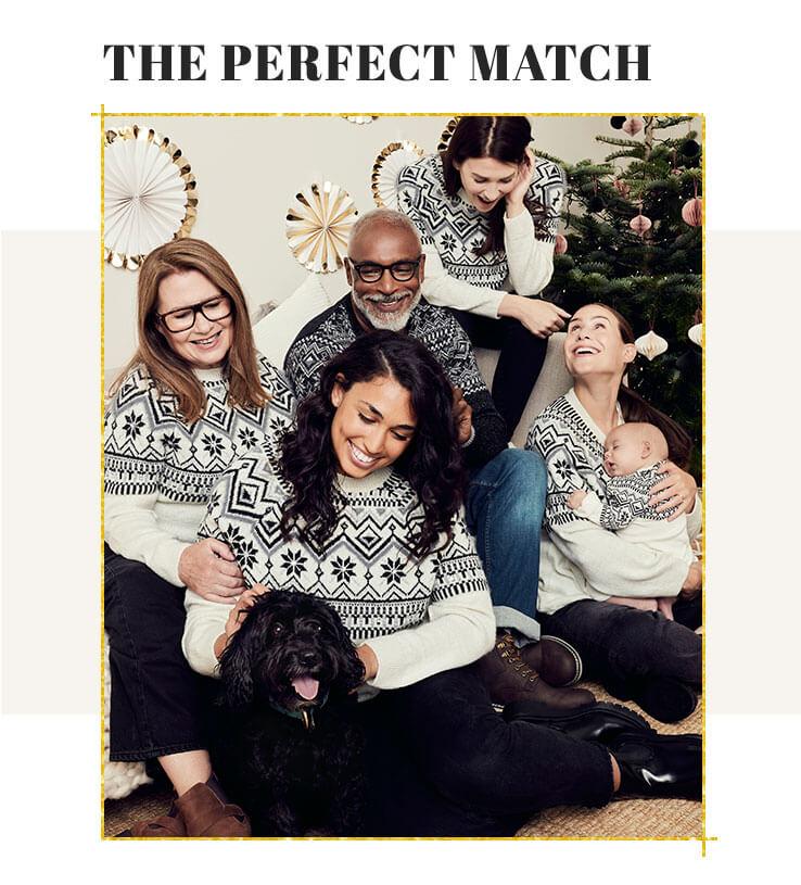 Four women, one man, a baby and a dog huddle together smiling on a sofa wearing matching white and navy Fairisle print knitted jumpers with Christmas tree and gold and white decorations hanging on the wall in the background.