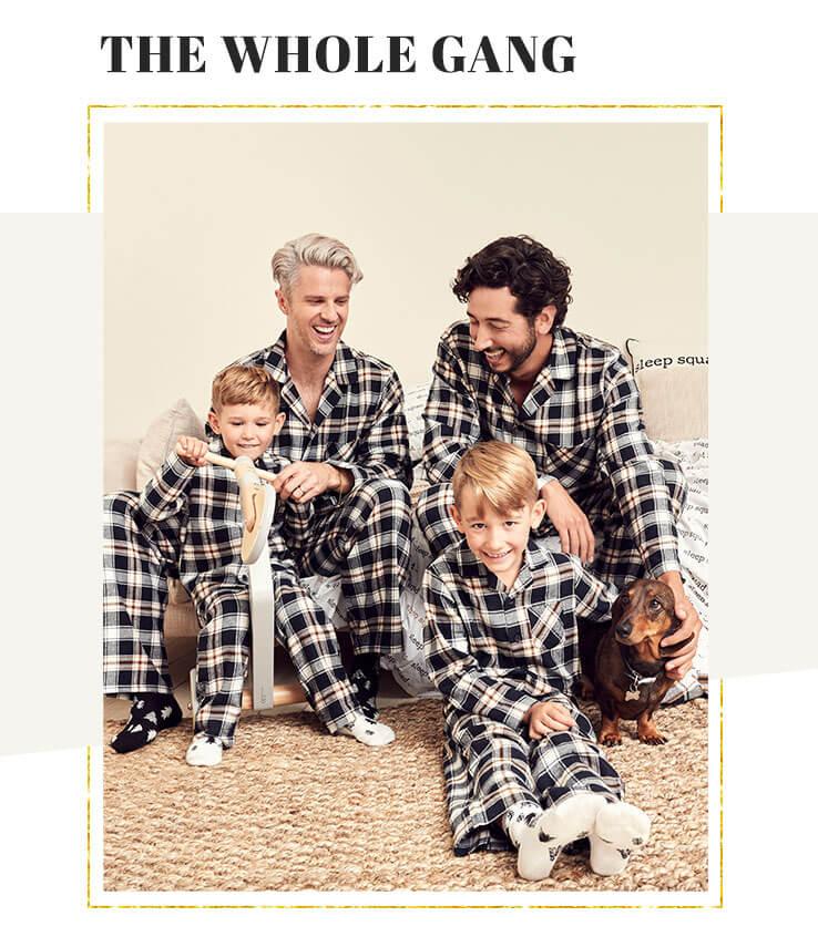 Two men, two young boys and a dog gather together on a cream sofa wearing blue and white check print family matching Christmas pyjamas and Christmas tree printed socks.