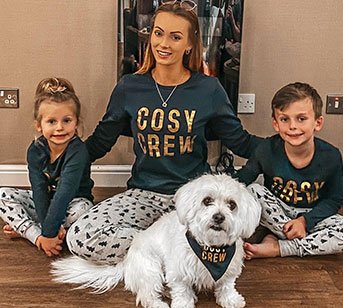 Young girl, woman, young boy and a dog all sit on a wooden floor wearing green cosy crew family matching Christmas pyjamas. 