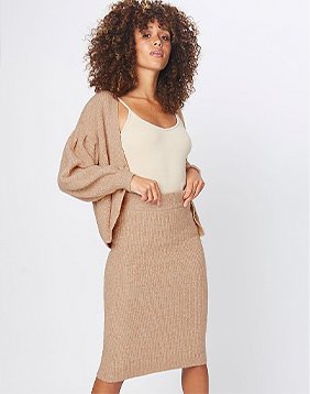 Woman with brown curly hair poses wearing brown knitted midi skirt, cream camisole and brown knitted cardigan.