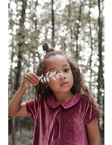 Girl in the woods covering her right eye with a leaf wearing a burgundy velour dress