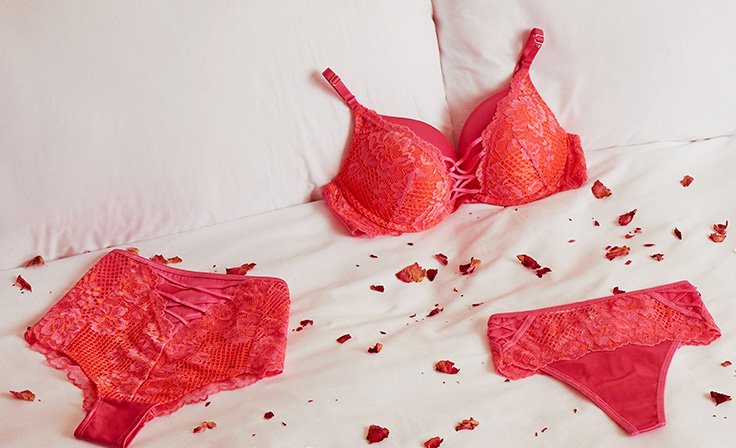 Red floral lace lingerie set laid on white bed with rose petal confetti.